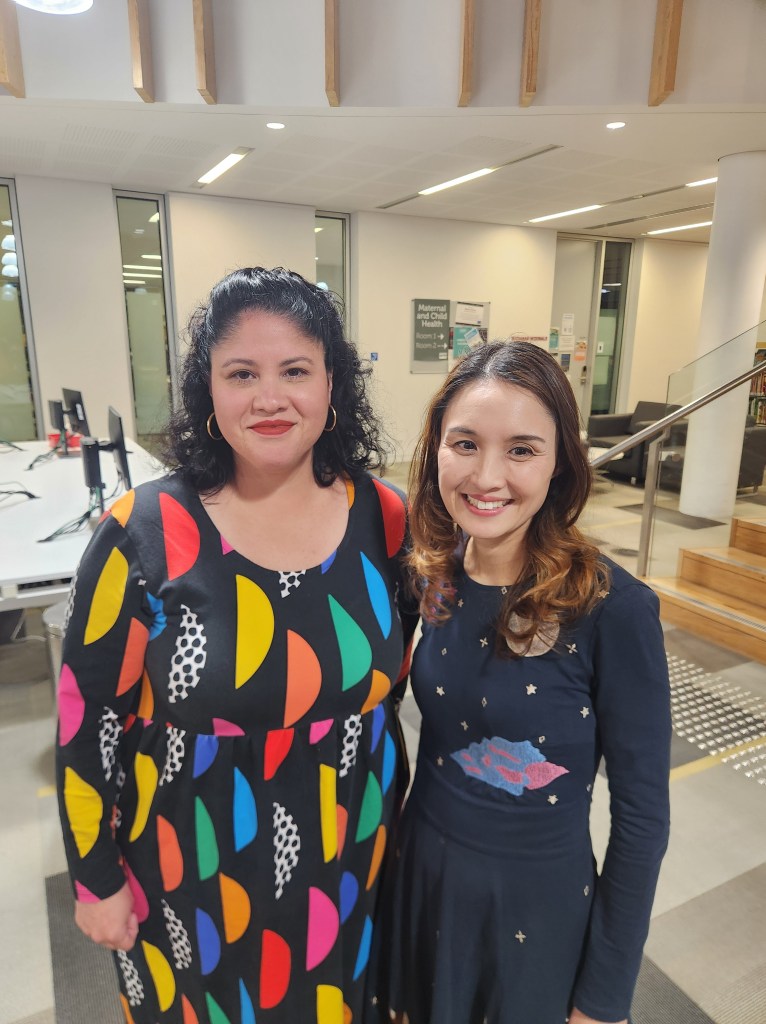 Photo of Zuleyka and Alice. Zuleyka is a Latin woman with dark curly hair who wears a colourful dress. Alice is a Chinese woman with light brown hair wearing a blue dress and she is smiling