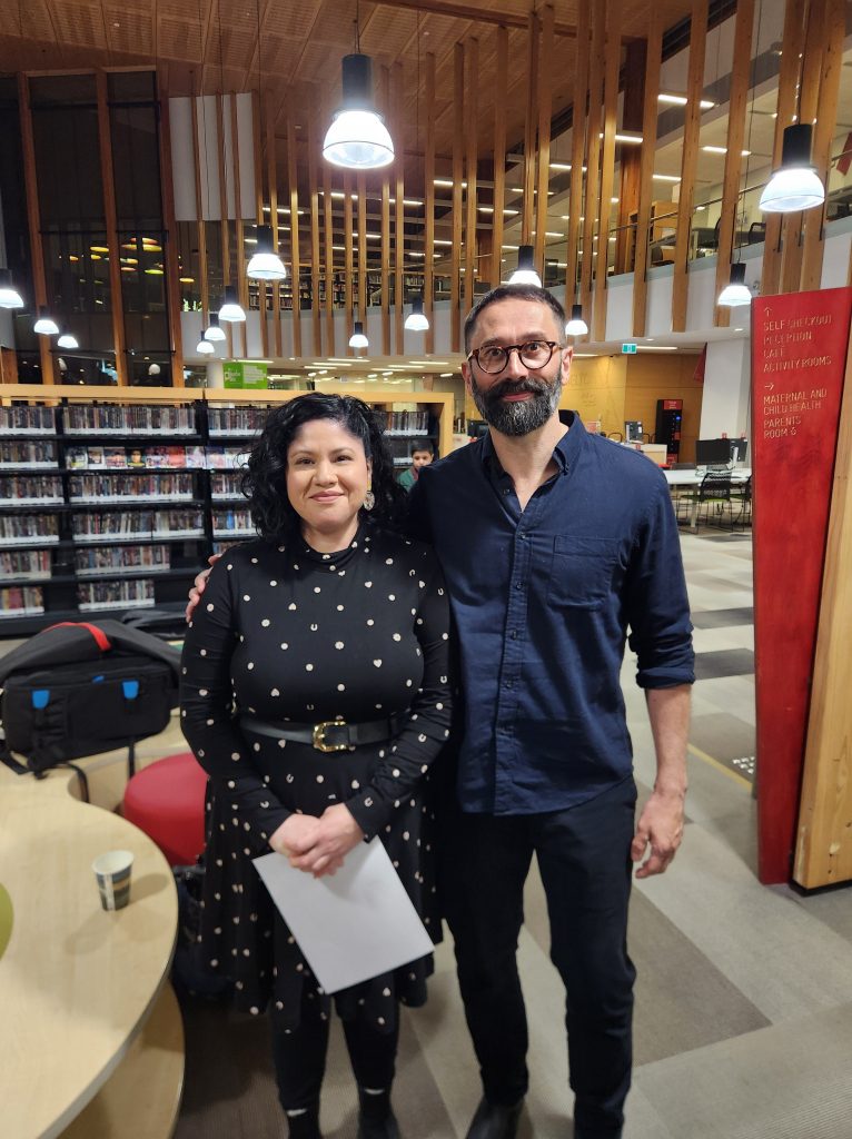 Zuleyka and Mark Brandi smile while posing in a library. Zuleyka is a Peruvian woman with short black curly hair, wearing a high-neck black dress with a gold print, holding a piece of paper. Mark has his arm around Zuleyka's shoulder. He is an Italian man with short salt and pepper hair and beard, who wears glasses and a long navy shirt and pants.