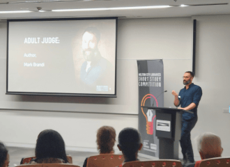 Mark Brandi holds his hand out as he speaks behind a podium, before a crowd. He stands beside a large screen with his photo and title: Adult Judge: Author, Mark Brandi. Mark is an Italian man with short salt and pepper hair and beard, who wears glasses and a long navy shirt and pants.