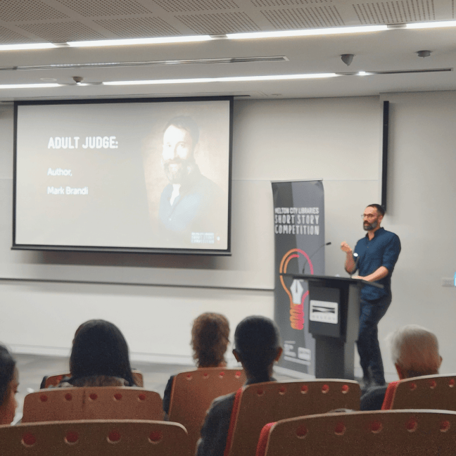 Mark Brandi holds his hand out as he speaks behind a podium, before a crowd. He stands beside a large screen with his photo and title: Adult Judge: Author, Mark Brandi. Mark is an Italian man with short salt and pepper hair and beard, who wears glasses and a long navy shirt and pants.